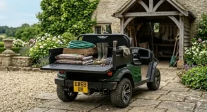 A two seat electric utility buggy with an open rear cargo bed loaded with gardening tools, wellies, and bags of organic compost.
