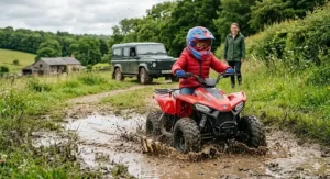 A child driving a red all terrain quad for kids through a muddy puddle on a countryside trail in the UK.