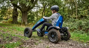 A rugged blue pedal go-kart with chunky pneumatic tyres being driven along a grassy, off-road woodland track.