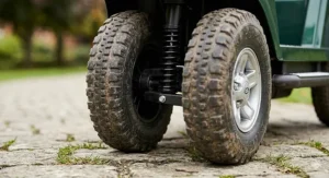 A front-angle detail view of a rugged, muddy, deep-treaded tyre and the complex metal and black plastic steering and suspension linkage on a green toy buggy.