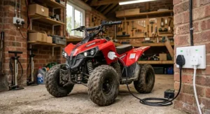 A close-up of an electric quad bike battery being charged using a standard UK three-pin plug in a garage workshop.
