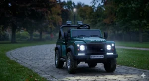 The front view of a kids' green ride-on buggy with Land Rover branding on a park path at dusk, with its LED headlights and running lights brightly illuminated.
