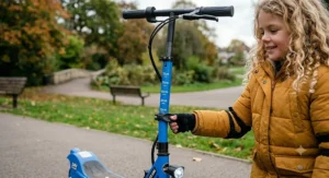 A photorealistic close-up illustration of the adjustable handlebar height settings on the blue 24v kids' electric scooter, with a child's hand using a clamp and visible text height markings in a park setting.