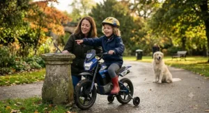 A young child on an electric motorbike with stabilisers pointing towards a garden feature in a park, with a parent and dog nearby.