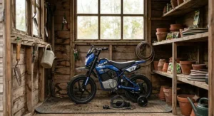 A children's electric motorbike with stabilisers parked inside a wooden garden shed with a battery charger visible on the floor.