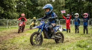 A child on a 24v electric dirt bike surrounded by friends holding a Union Jack, wearing full safety gear in a grass field.