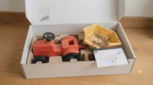 A flat lay view of a brand new ride-on tipping truck and trailer, partially unboxed from its cardboard packaging on a wooden floor, ready for quick assembly.
